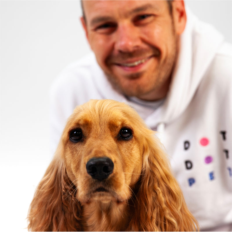 Man wearing a white hoodie with colorful letters, sitting next to a brown dog.